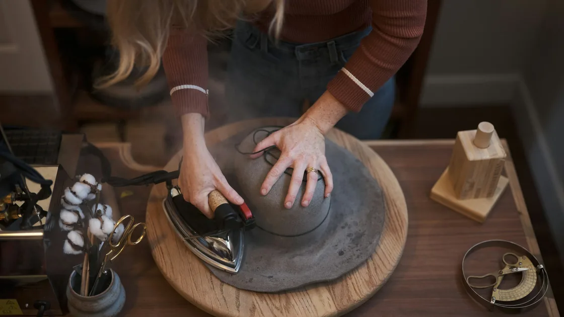 Manos alisando con una plancha una copa de fieltro colocada sobre una horma de madera, entre vapor, sobre un banco de taller con herramientas de sombrerería